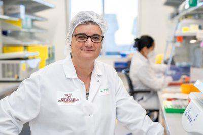 A woman in a white lab coat and with a hair net stands in her medical laboratory.