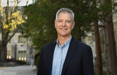 Pitts smiles outside with trees and stone buildings in background