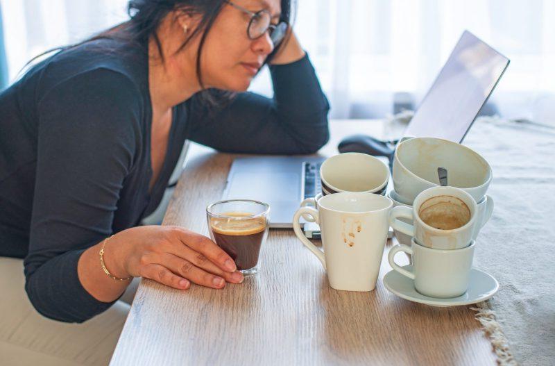 Woman sits in front of a computer with a cup of coffee with several empty coffee cups nearby 